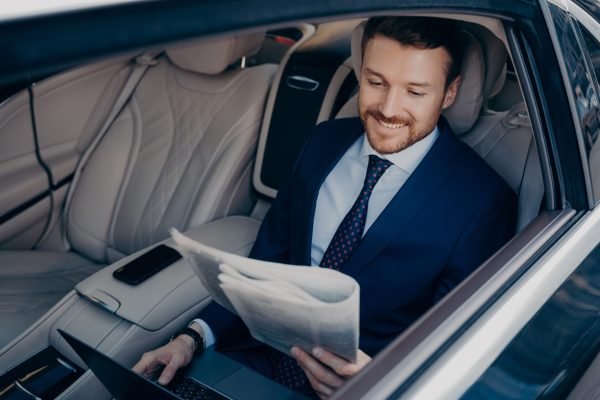 Relaxed entrepreneur reading newspaper while riding in limousine Relaxed male entrepreneur dressed in blue suit, reading latest newspaper and checking news regarding his successful company, while working on notebook, taking ride to work